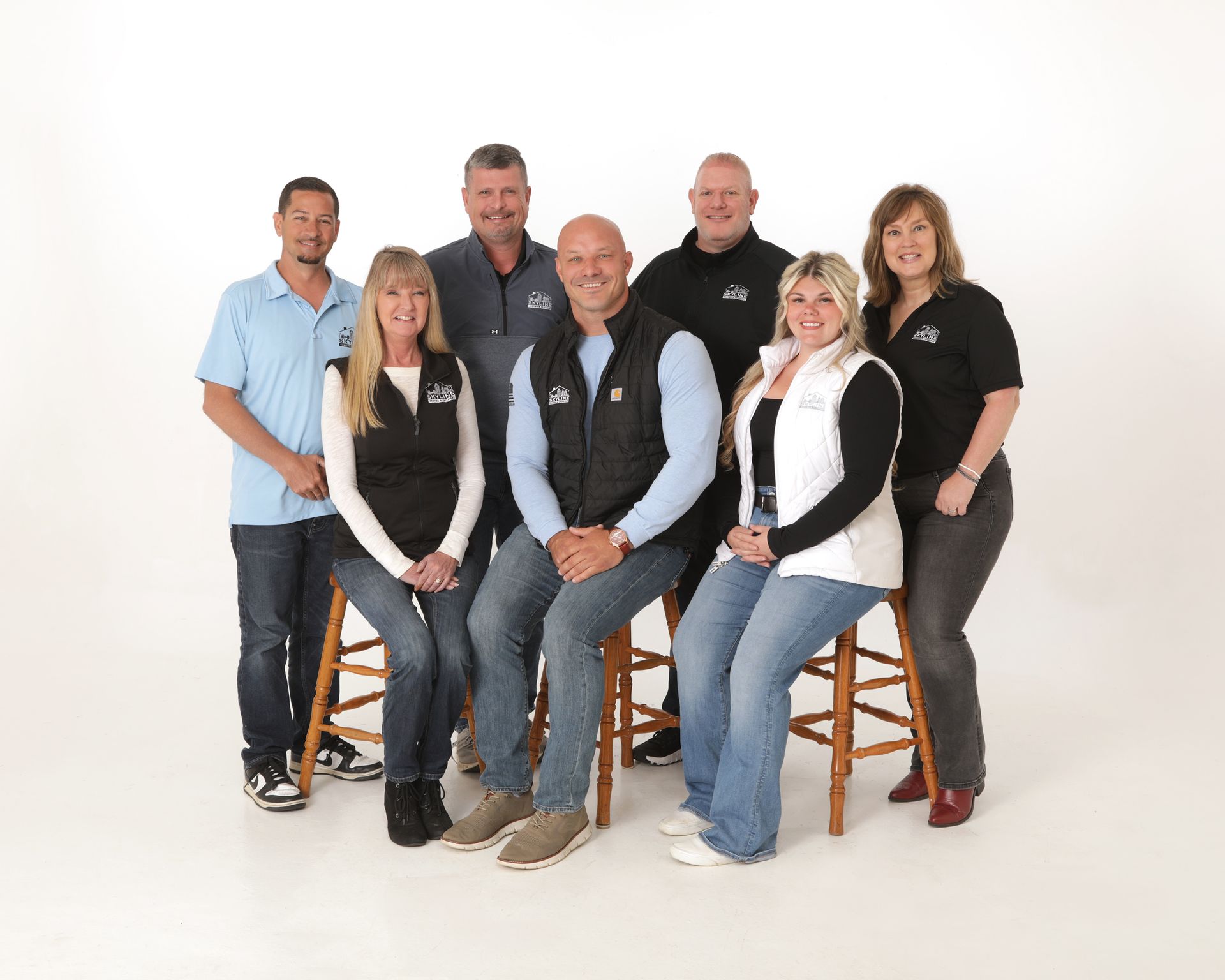 A group of seven smiling professionals posing together in a studio setting with wooden stools against a white background.